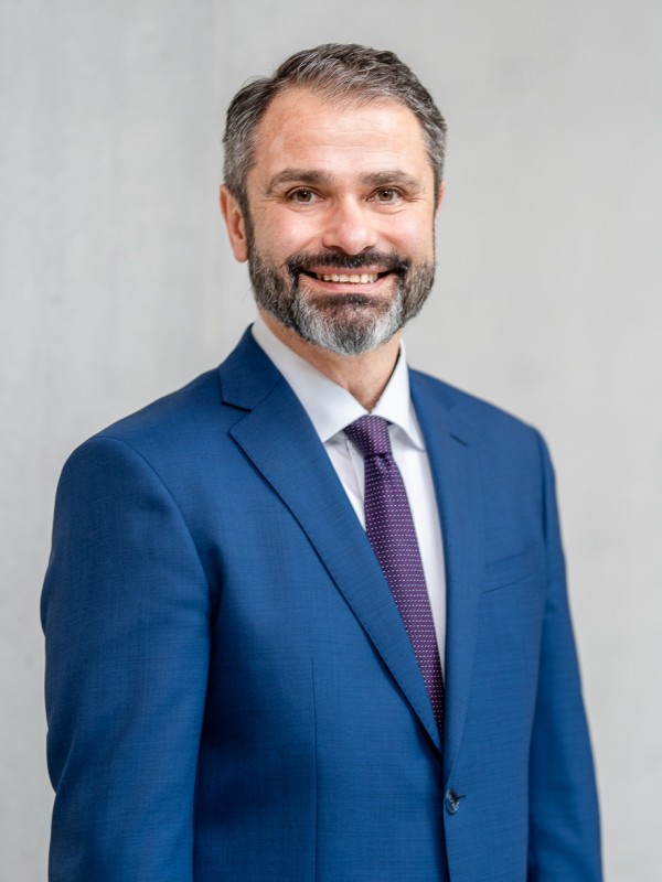 Portrait von Herrn Hauser, lächelnd in blauem Anzug mit Krawatte