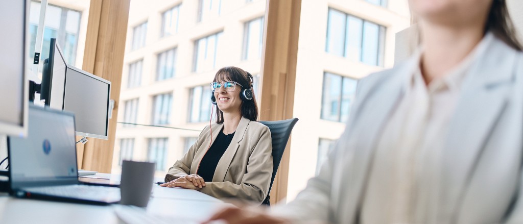 Frau mit Headset und Brille telefoniert am Arbeitsplatz und lächelt freundlich