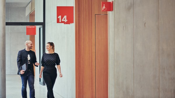 Eine junge Frau mit einem schwarzen Pullover mit drauf genähten Perlen und ein junger Mann in dunklem Jackett und Jeans unterhalten sich während sie durch die Gänge des Campus laufen