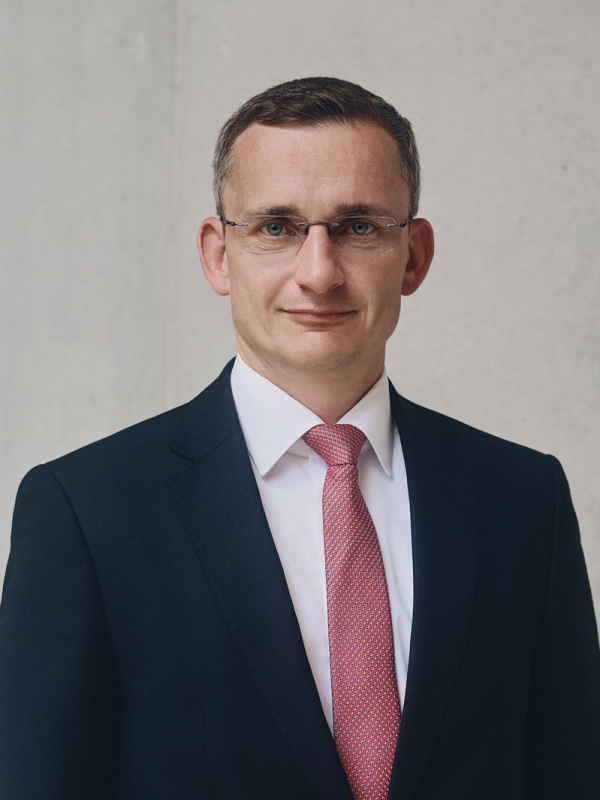 Portrait von Matthias Bogk, Chief Financial Officer, Chief Risk Officer und Vorstandsmitglied lächelt im Anzug vor grauer betonwand.
