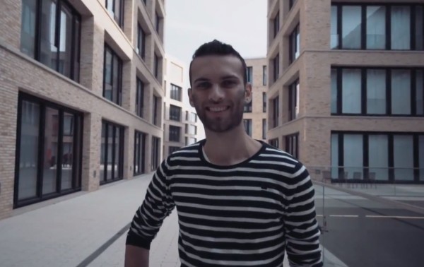 Junger Mann steht lächelnd im Campus-Hof und schaut in die Kamera