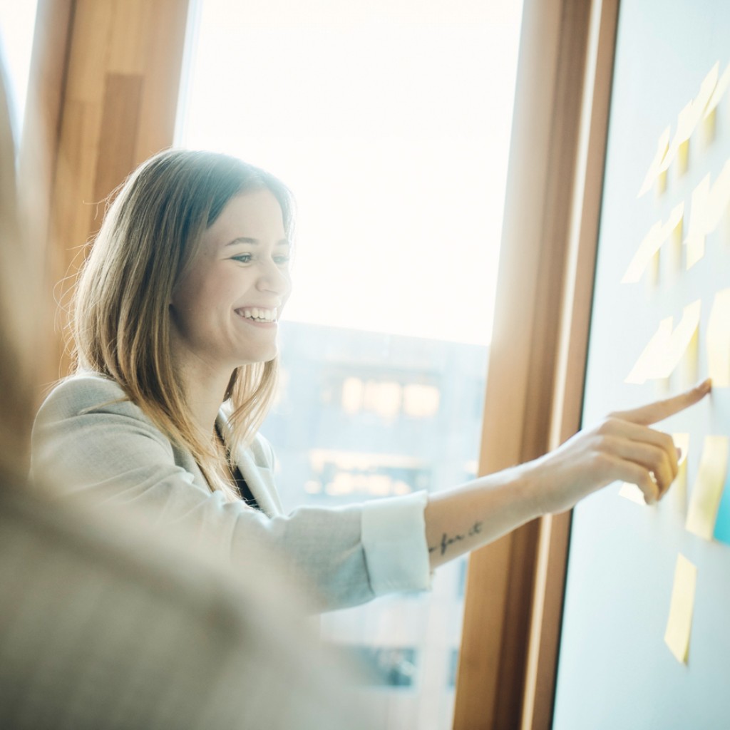 Mitarbeiterin-zeigt-auf-beschriebene-Post-Its-an-der-Wand