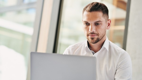 Ein junger Mann in weißem Hemd der auf einen silbernen Laptop schaut