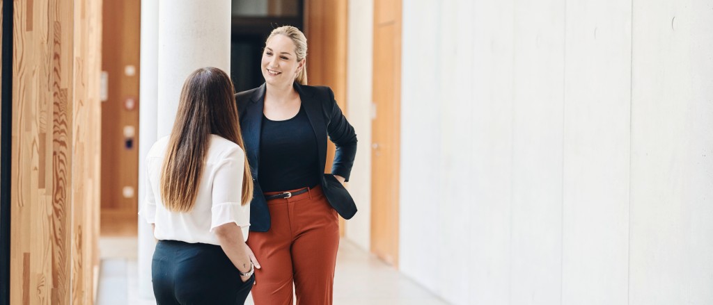 Eine kleine dunkelhaarige und eine blonde große Frau die sich in einem Gang unterhalten