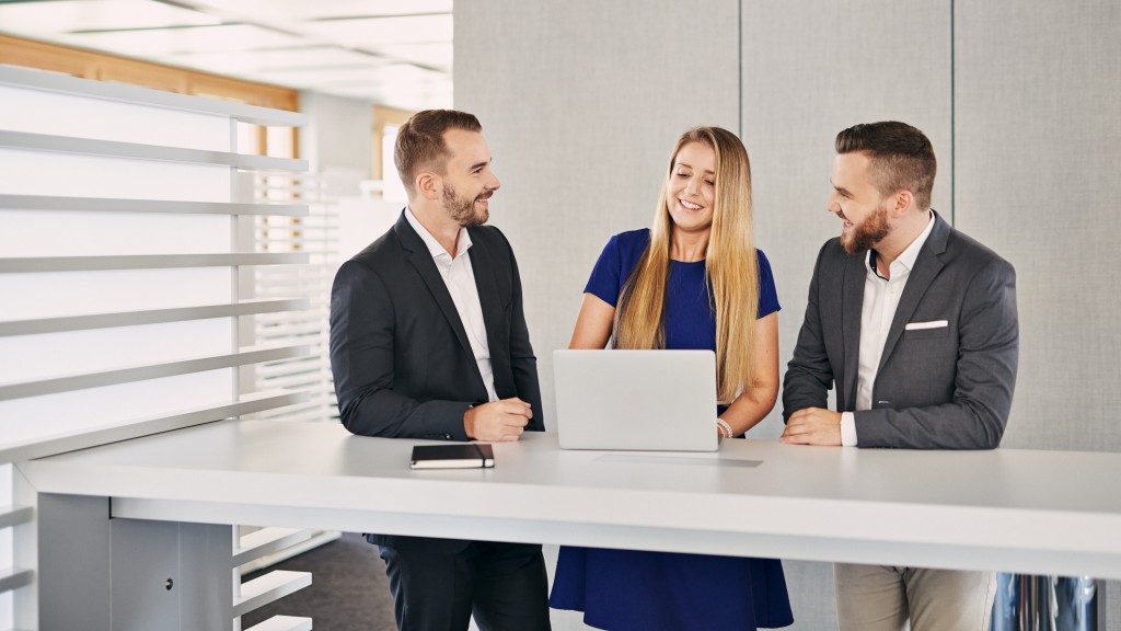 Zwei junge Männer in dunklen Anzügen die einer blonden Frau mit blauem Kleid zwischen ihnen zuhören während sie etwas von einem silbernem Laptop abliest