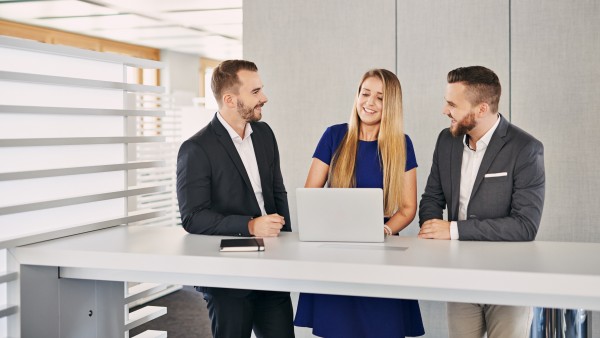 Zwei junge Männer in dunklen Anzügen die einer blonden Frau mit blauem Kleid zwischen ihnen zuhören während sie etwas von einem silbernem Laptop abliest