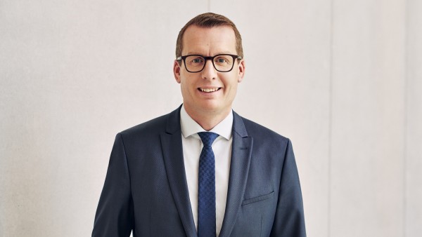 Freundlicher Mann mit Brille steht im Anzug mit Krawatte vor grauer Betonwand