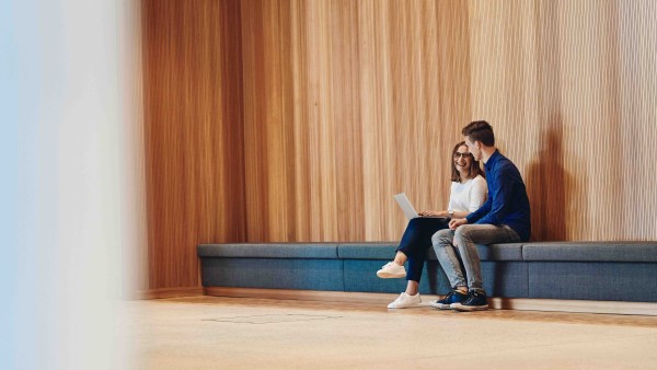 Zwei junge Mitarbeiter sitzen auf einer Sitzgelegenheit und arbeiten gemeinsam an einem Laptop