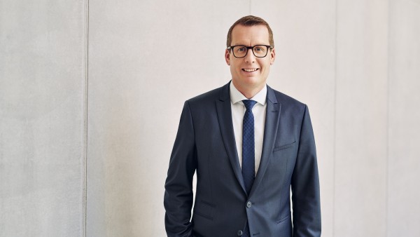 Freundlicher Mann mit Brille steht im Anzug mit Krawatte vor grauer Betonwand
