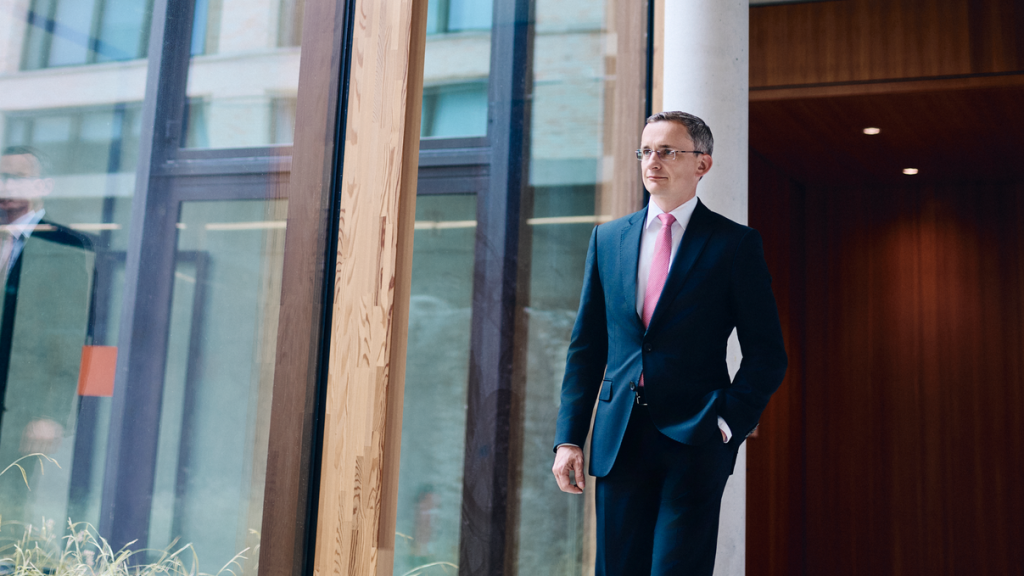 Freundlicher Herr im Anzug mit Brille, läuft einen lichtdurchfluteten Gang des W&W Campus entlang